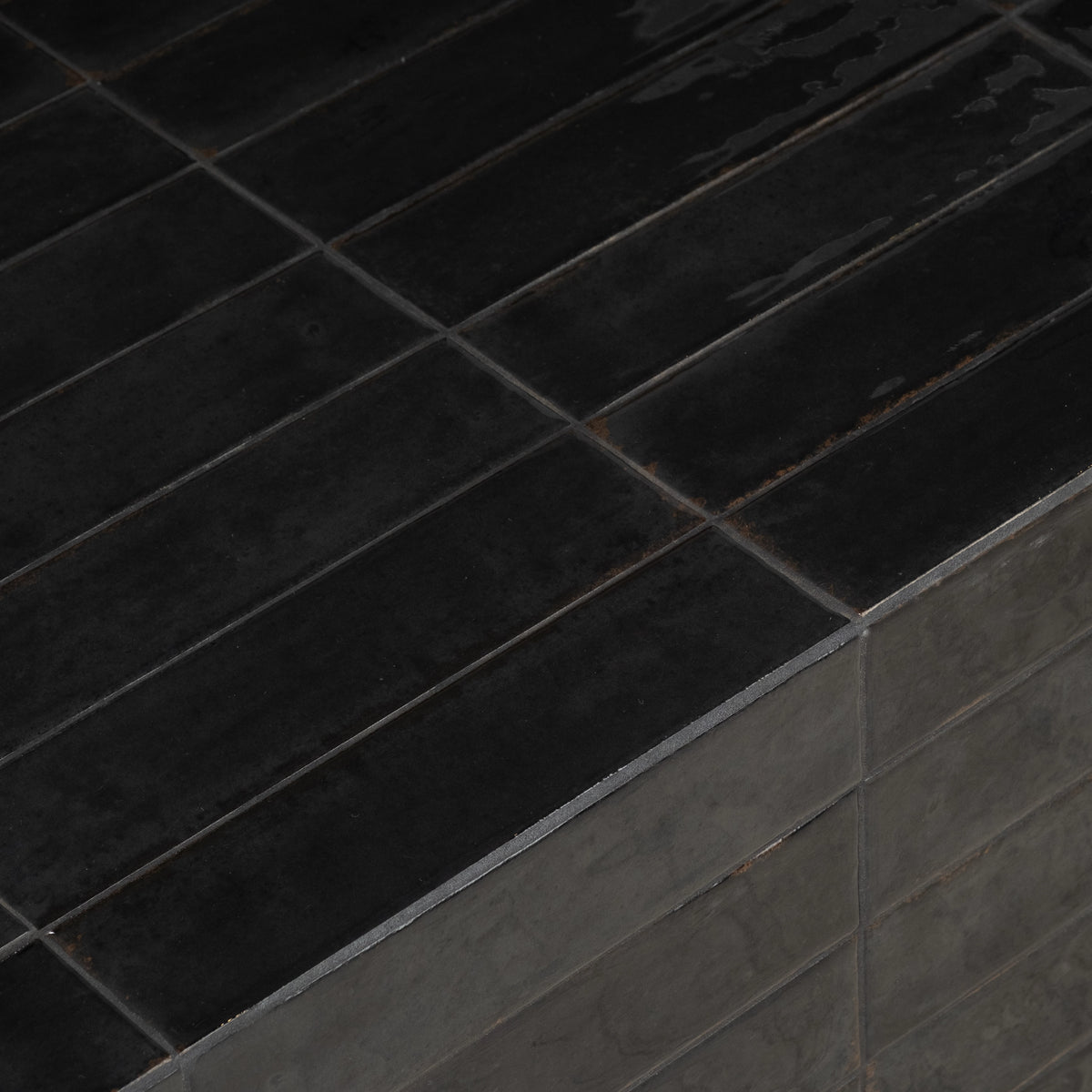 Close-up showing the corner of the Bloor Coffee Table in Black glazed tile, with the glossy tabletop strips, thin grout lines, and softer charcoal side tiles below.