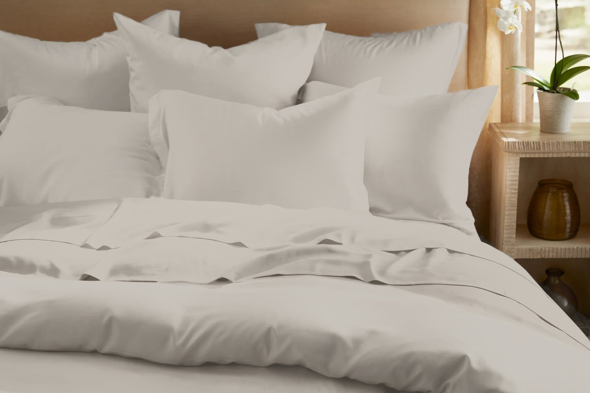 Front view of the Legna Classic Bedding Set in Cappuccino, a warm greige satin-finish weave, showing stacked euro pillows, tailored flanged pillowcases, and a flat sheet turned over a smooth duvet cover.