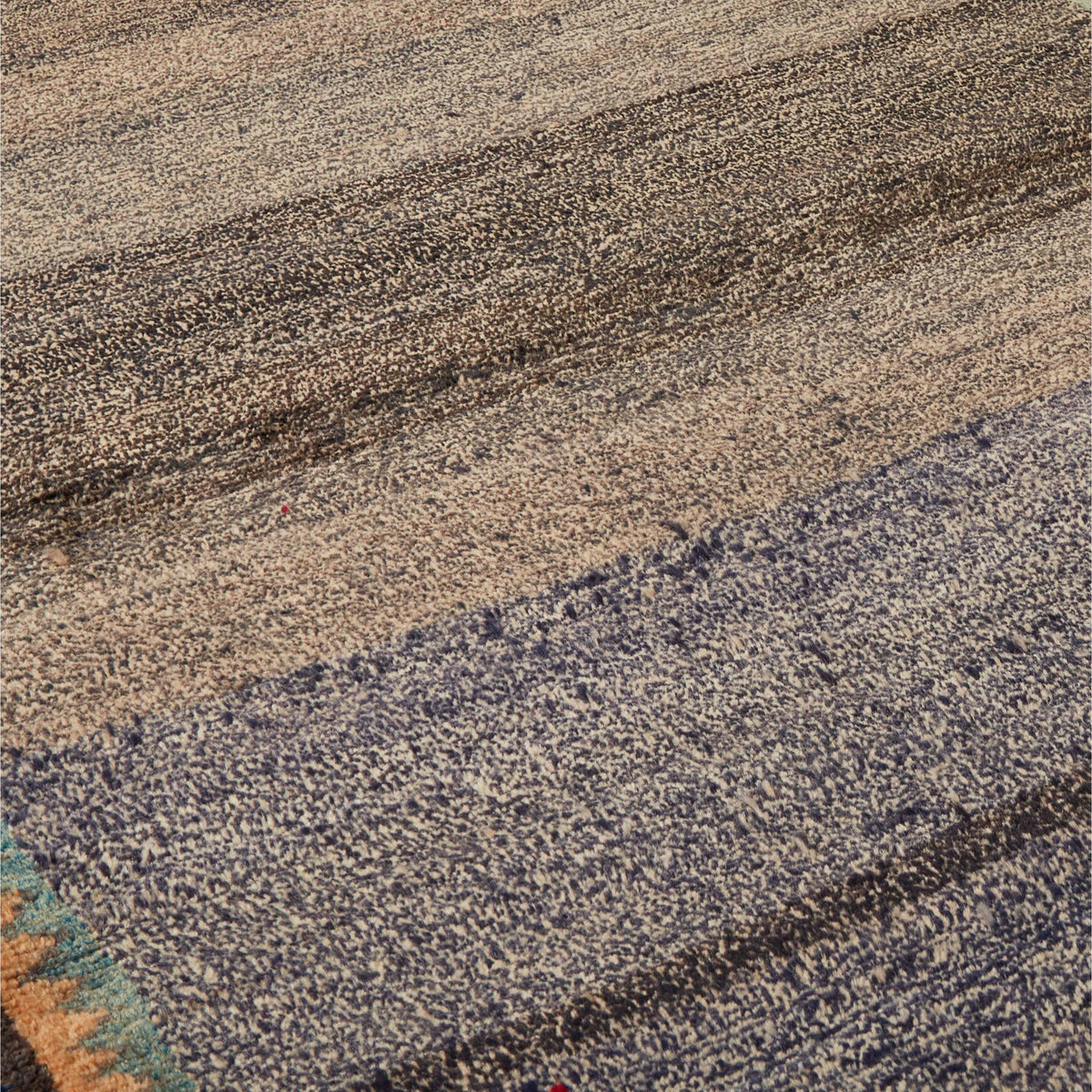 Close-up of a textured area rug with a modern abstract design, featuring horizontal bands of beige, brown, and slate blue with a small multicolor accent at the corner.
