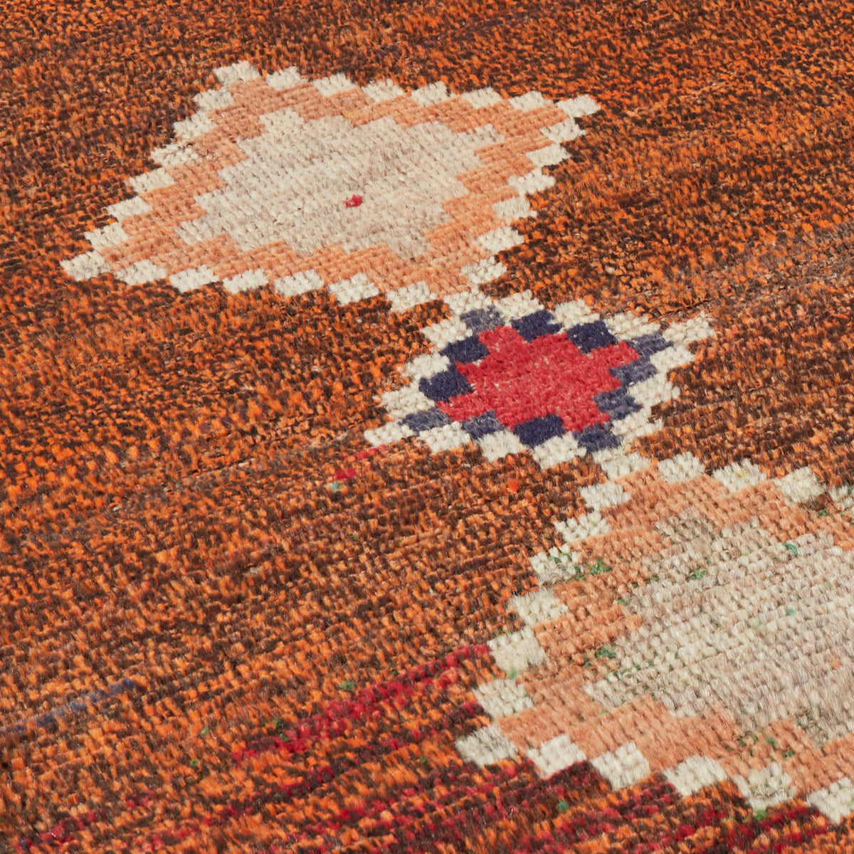 Close-up of a textured woven rug with a rust orange field and geometric motifs in cream, peach, red, and dark blue.