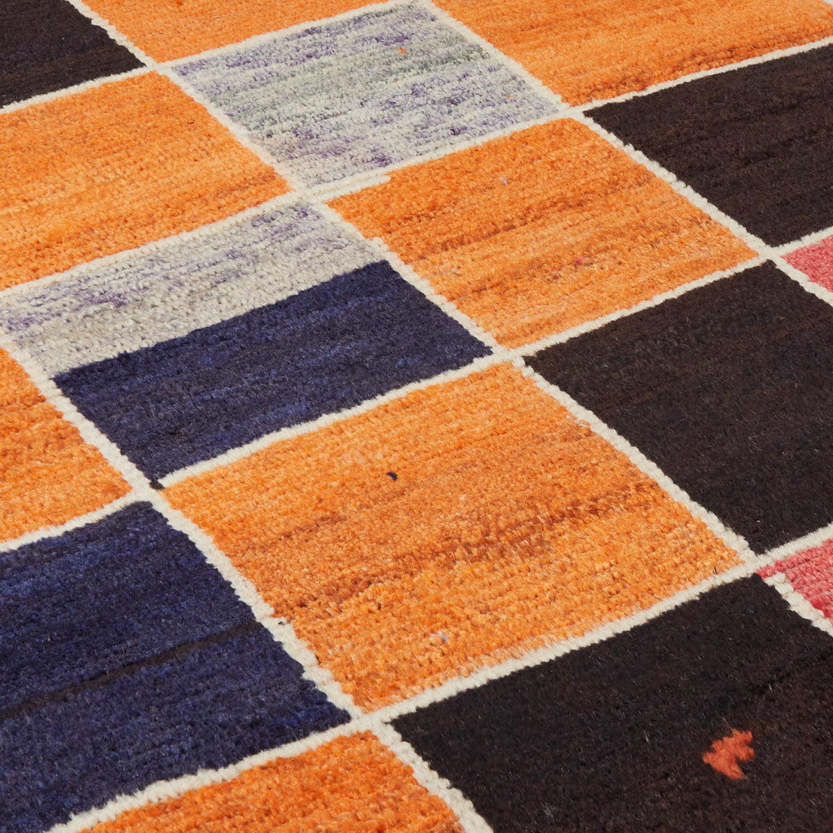 A close-up of a woven geometric area rug featuring a patchwork design of orange, dark brown, navy blue, and light gray squares separated by off-white borders.