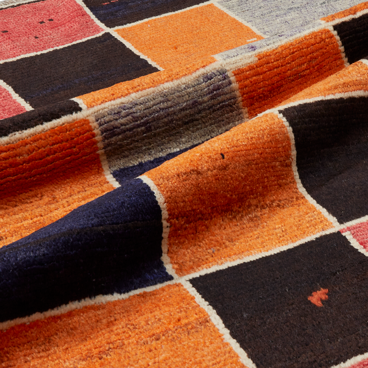 A close-up of a textured wool area rug with a geometric patchwork design in orange, black, navy blue, gray, and muted red, separated by cream-colored borders.