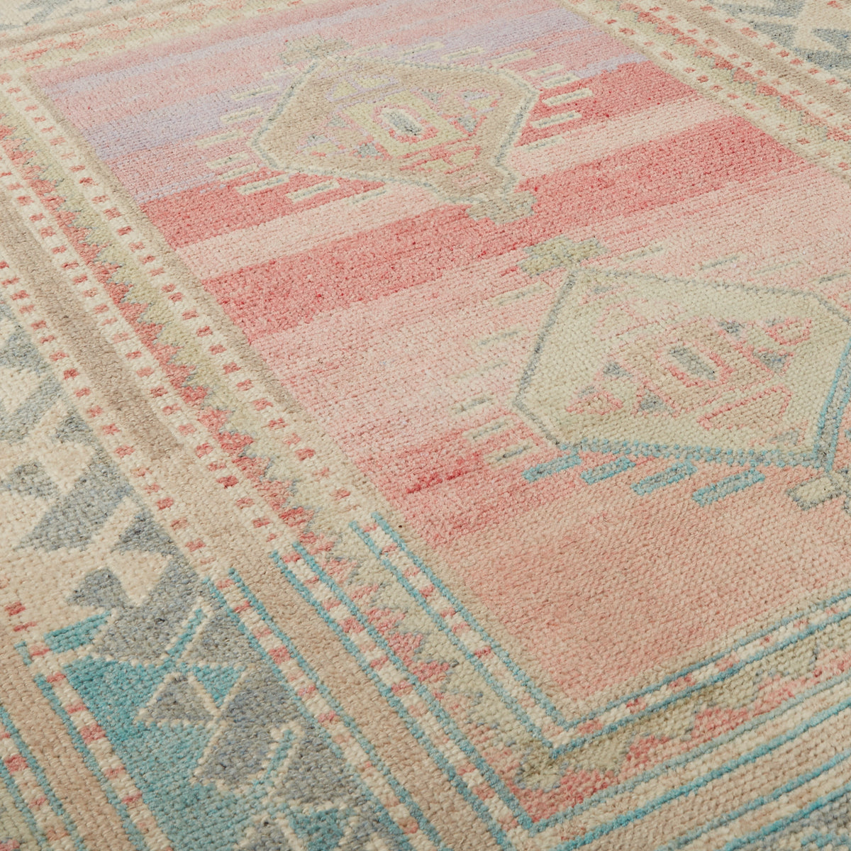 A close-up of a woven rug with a distressed geometric pattern in soft pink, beige, light blue, and gray.