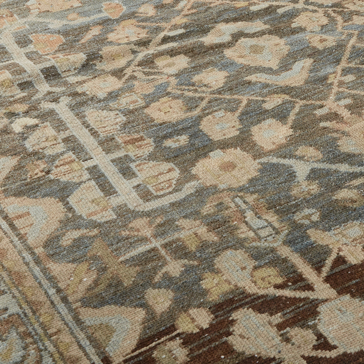 A close-up of a traditional patterned rug with a muted blue-gray background, beige and cream floral motifs, and brown accents.