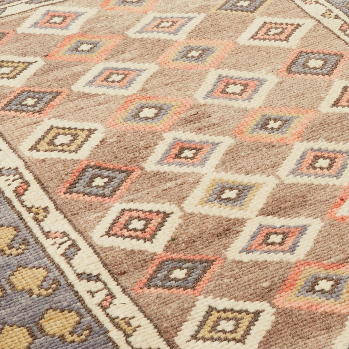 A close-up of a woven area rug with a geometric diamond pattern in beige, cream, gray, muted orange, and brown tones, showing the detailed texture and corner design.