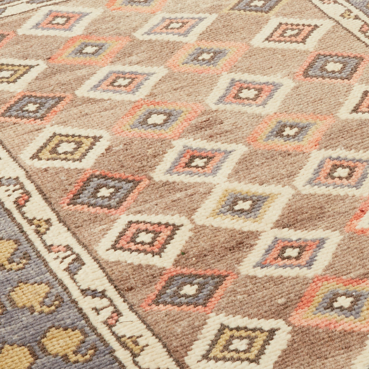 A close-up of a woven rug with a geometric diamond pattern in beige, cream, gray, and muted coral tones, showing the texture and part of the border.
