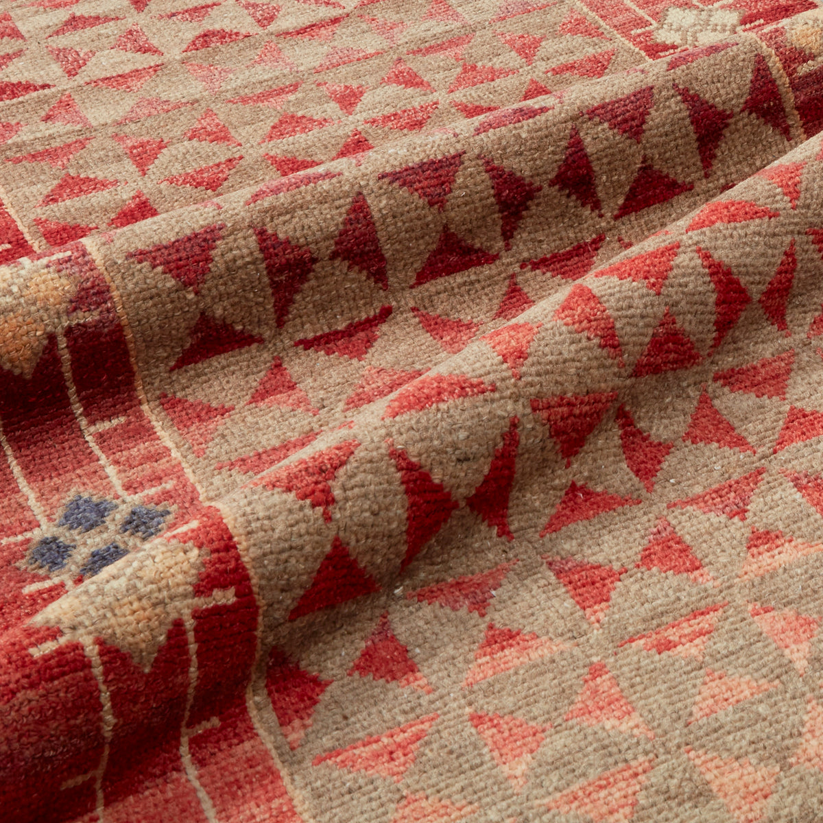 A close-up of a woven textile with a geometric pattern in red, pink, and beige, showing the texture and folds of the fabric.